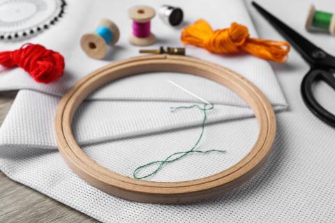 Supplies for needlepoint including wooden embroidery hoop, thread in various colors, black scissors, white fabric, and an embroidery needle with green thread.