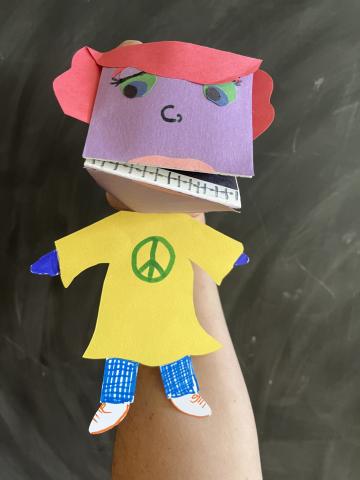 A paper puppet with pink hair, purple face, braces on the teeth, yellow shirt, and blue pants.