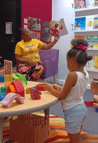 Author, Dayonne Necole, reading a book aloud with a child playing and listening.